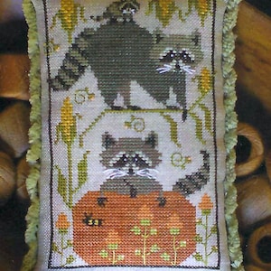 May include: Cross-stitch pillow with a green fringe. The design features two raccoons, one atop a pumpkin and the other above. Corn stalks and other foliage surround the raccoons. The color palette includes shades of green, orange, and tan.