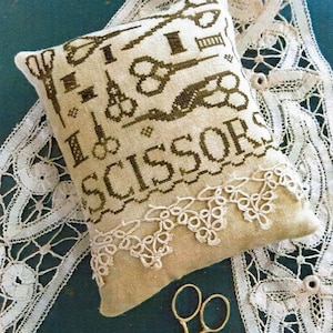 May include: A beige needlepoint pillow with a green embroidered design of scissors and the text "I Love Scissors". The pillow is trimmed with white lace and is sitting on a white lace doily.