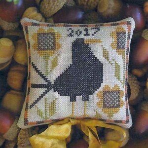 May include: A cross-stitch pillow with a black bird design, surrounded by sunflowers and leaves. The pillow has a brown and white plaid border and a yellow ribbon tied around it. The text "Fall 2017" is stitched in the center of the pillow.