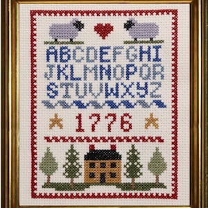 May include: A white cross-stitch sampler with a red border. The sampler features a blue alphabet, two gray sheep, a red heart, a row of stars, the number 1776, and a small house with trees.