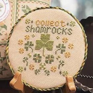 Puede incluir: Un diseño circular de punto de cruz con las palabras "collect shamrocks" y motivos de tréboles en verde y amarillo. El diseño está sobre una tela beige, enmarcado con un cordón trenzado verde y amarillo. Se presenta sobre un pequeño soporte de madera.