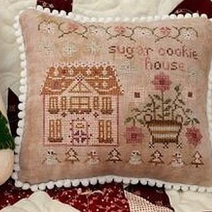 May include: A decorative pillow with a cross-stitch design. The pillow features a gingerbread house, flowers, and the text "sugar cookie house." The pillow has a light pink background and white pom-pom trim.