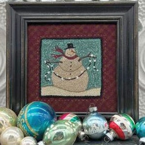 May include: A framed, stitched artwork of a snowman wearing a black top hat and red scarf, holding a string of lights. The artwork is set against a burgundy background and framed in a dark wood. Christmas ornaments in various colors are in the foreground.