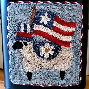 May include: A patriotic, hand-stitched rug featuring a sheep pulling a cart with an American flag. The rug has a blue background with red, white, and blue accents. The border is trimmed with a red, white, and blue rope.