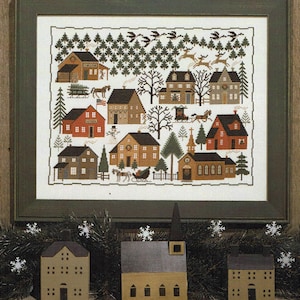 May include: A cross-stitch pattern of a winter village scene with houses, a church, trees, and reindeer. The pattern is framed in a dark green frame and is displayed on a wooden surface with three miniature houses.