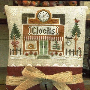 May include: A beige pillow with a cross-stitch design of a clock shop. The shop has a red roof, a brown door, and a sign that says "Clocks". The pillow is tied with a brown ribbon and a bell.