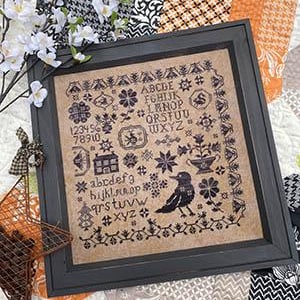 Puede incluir: Un sampler de punto de cruz enmarcado con un diseño en blanco y negro que presenta un cuervo, una casa, flores y el alfabeto. El sampler está sobre un fondo blanco con tela estampada en blanco y negro.