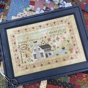 以下が含まれることがあります： 青い車、木、花のある家のクロスステッチ刺繍。アルファベットは花柄のデザインで枠の周りに刺繍されています。刺繍は濃い青色の額縁に入っています。