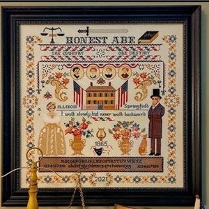 Puede incluir: Un sampler de punto de cruz enmarcado con el texto "HONEST ABE" y "ONE COUNTRY ONE DESTINY." El diseño incluye ilustraciones de un edificio, figuras, banderas y arreglos florales. También está presente el texto "ILLINOIS Springfield" y "I walk slowly but never walk backward". El año 2021 está en la parte inferior.