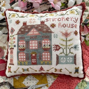 Puede incluir: Un cojín cuadrado decorativo con un diseño de punto de cruz. El cojín presenta una casa con el texto "stitchery house" y detalles florales. La paleta de colores incluye tonos de azul, rojo y crema, con un borde con volantes.