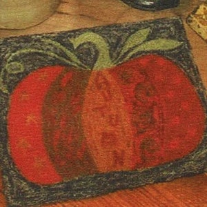 May include: A close-up of a framed needlepoint design featuring a red, orange, and brown pumpkin with green leaves. The background is a dark brown.