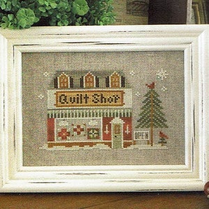 May include: Framed cross-stitch art depicting a "Quilt Shop" with a red and white facade, a green Christmas tree, and snowflakes. The shop sign is brown with white lettering. The frame is white with a distressed finish.