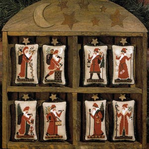 May include: Eight embroidered Santa Claus ornaments in a wooden display case. Each ornament features a different Santa Claus design with a red and white color scheme. The ornaments are displayed in a wooden case with a crescent moon and stars.