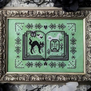 May include: Cross-stitch artwork in a decorative silver frame. The image shows a green background with a black cat, a ghost, and "RIP" on an open book. The frame has an ornate design, ideal for Halloween decorations.