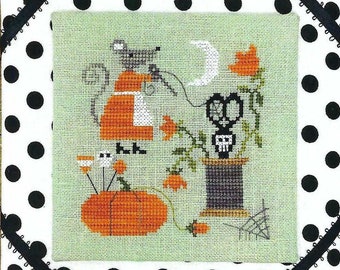 May include: A cross-stitch pattern featuring a black cat, a pumpkin, and a spool of thread with a skull-shaped scissors. The design is set against a green background with a white polka dot border.
