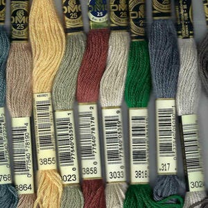 May include: A collection of DMC embroidery floss in various colors, including blue, beige, yellow, green, red, and gray. Each skein is labeled with a number and barcode. The floss is tightly wound and ready for use in needlework projects.