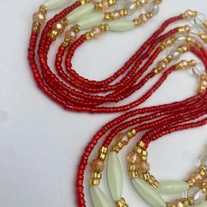 May include: Close-up of multiple strands of beaded jewellery. The strands feature red, gold, clear, and pale green beads. The beads are arranged in a layered design, creating an intricate pattern.