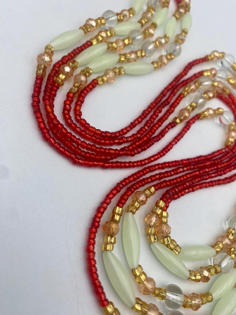 May include: Close-up of multiple strands of beaded jewellery. The strands feature red, gold, clear, and pale green beads. The beads are arranged in a layered design, creating an intricate pattern.