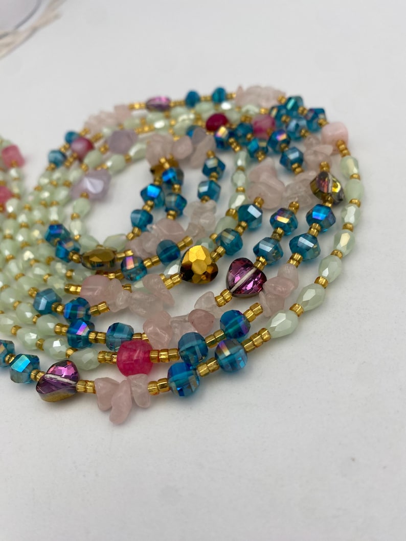 May include: A collection of beaded necklaces featuring a variety of colorful beads. The necklaces include iridescent blue, pink, and gold heart-shaped beads, and light green and pink stone beads. The beads are strung on gold-colored wire.
