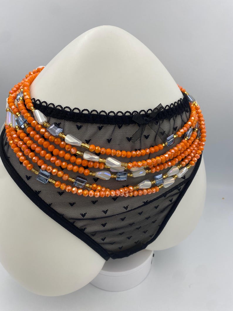 May include: A black lace lingerie set with a beaded waist chain. The waist chain features orange, blue, and white beads. The lingerie is displayed on a white mannequin.