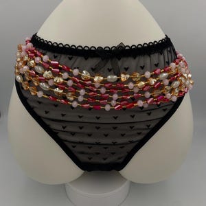 May include: Black lace underwear with a heart pattern, decorated with strands of pink, red, and gold beads. The underwear has a scalloped edge and a small bow.