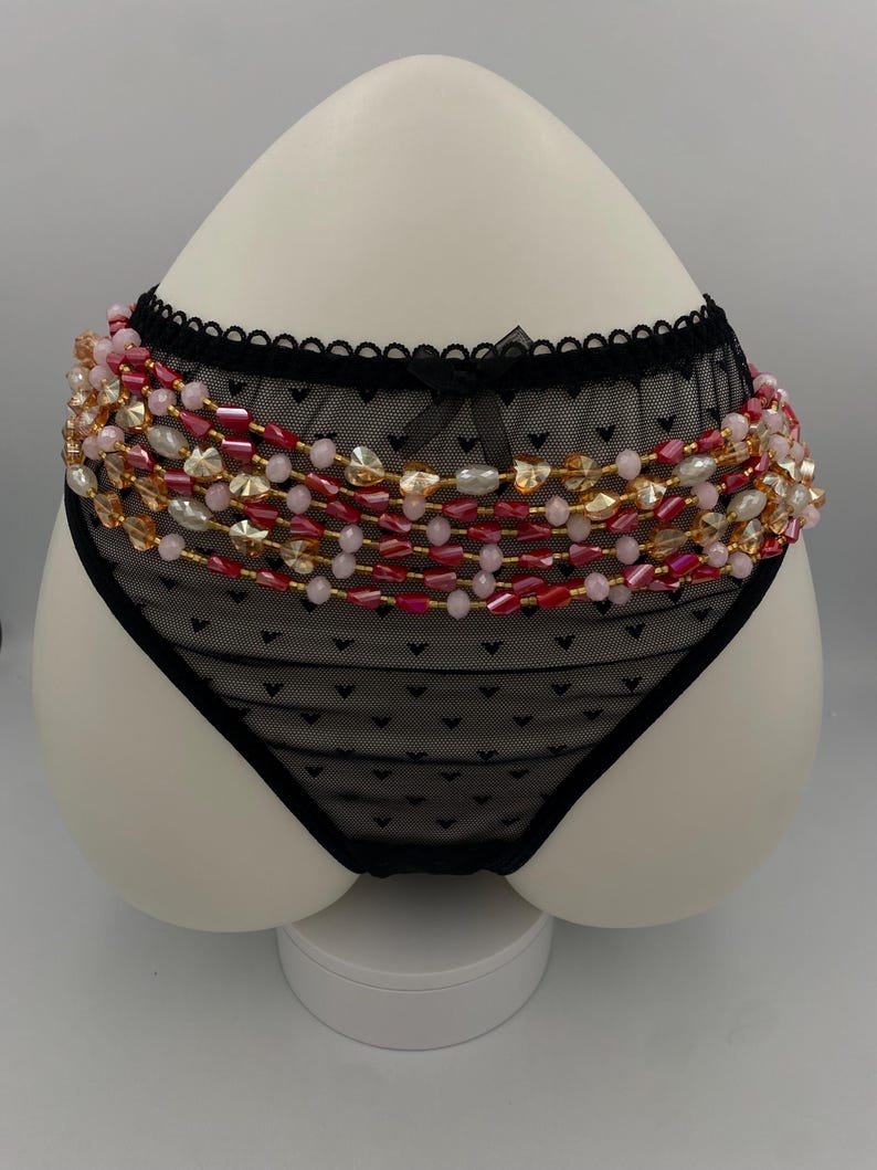 May include: Black lace underwear with a heart pattern, decorated with strands of pink, red, and gold beads. The underwear has a scalloped edge and a small bow.