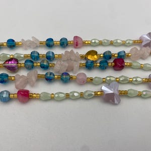 May include: Close-up of several beaded necklaces. The necklaces feature a variety of beads in shades of pink, blue, and gold. Some beads are faceted, while others are smooth. The necklaces are strung on gold-colored wire.
