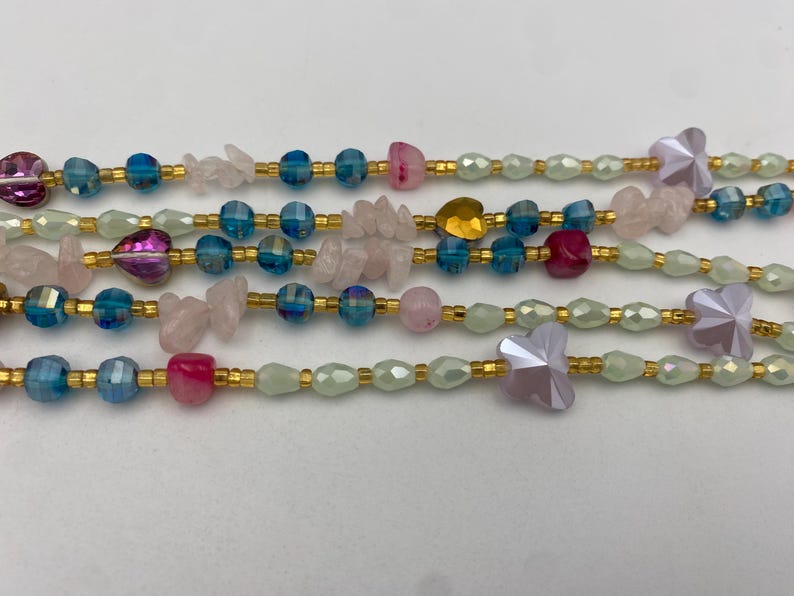 May include: Close-up of several beaded necklaces. The necklaces feature a variety of beads in shades of pink, blue, and gold. Some beads are faceted, while others are smooth. The necklaces are strung on gold-colored wire.