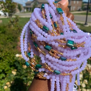 May include: A set of purple and gold waist beads with green accents. The beads are strung on a thin cord and are arranged in a spiral pattern. The beads are made of glass and have a smooth, polished finish.