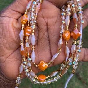 May include: A collection of beaded waist beads in shades of orange, gold, and clear. The beads are strung together with gold-coloured string. The beads are a mix of shapes, including round, faceted, and flower-shaped beads.