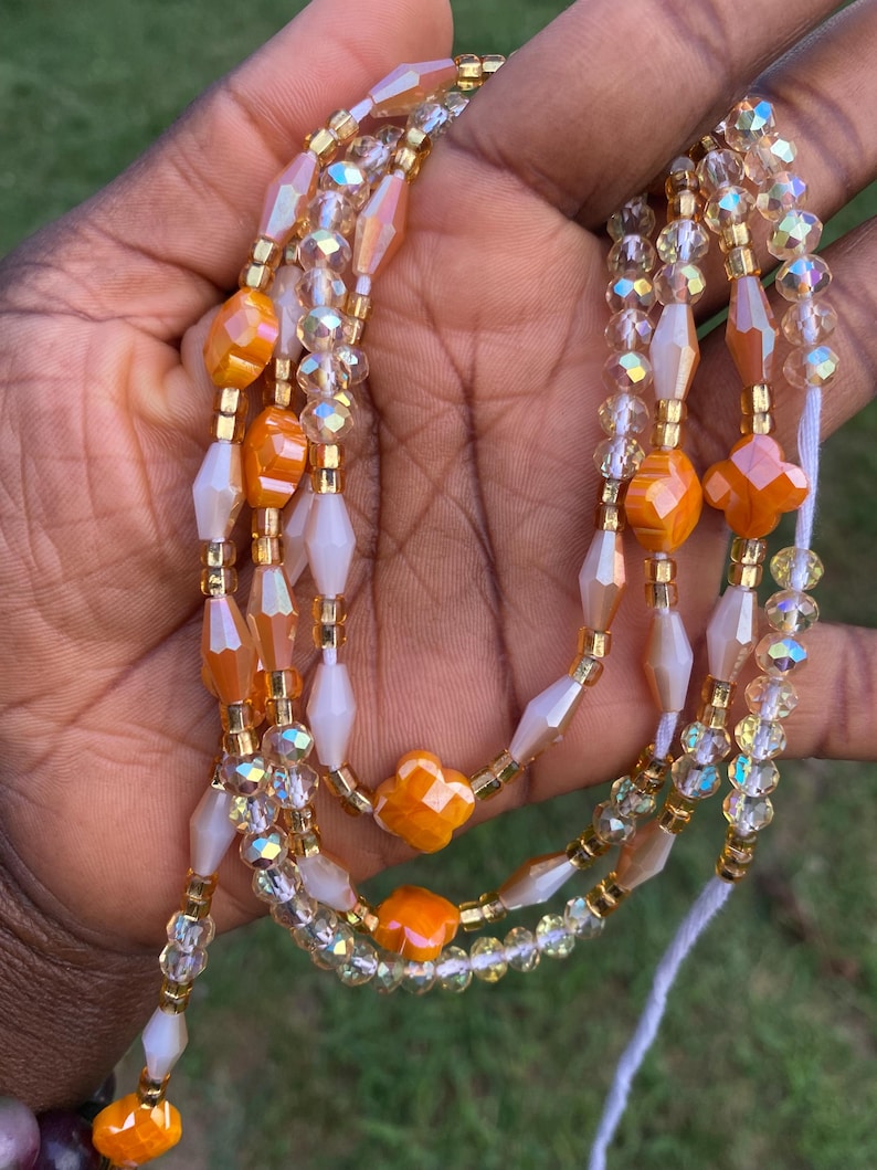 May include: A collection of beaded waist beads in shades of orange, gold, and clear. The beads are strung together with gold-coloured string. The beads are a mix of shapes, including round, faceted, and flower-shaped beads.
