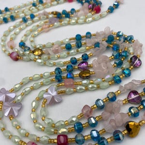 May include: Multiple strands of beaded necklaces in various colors. The necklaces feature a mix of light green, blue, pink, and gold beads. Some strands have flower-shaped beads and other unique shapes.