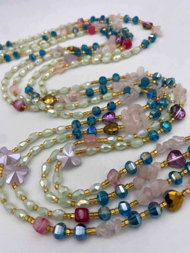 May include: Multiple strands of beaded necklaces in various colors. The necklaces feature a mix of light green, blue, pink, and gold beads. Some strands have flower-shaped beads and other unique shapes.