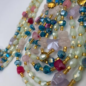 May include: Close-up of several beaded necklaces in shades of green, blue, pink, and purple. The beads are various shapes and sizes, including faceted crystals and irregular stones, strung with gold-colored spacers.