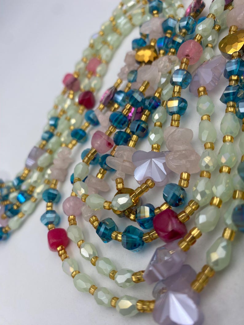 May include: Close-up of several beaded necklaces in shades of green, blue, pink, and purple. The beads are various shapes and sizes, including faceted crystals and irregular stones, strung with gold-colored spacers.