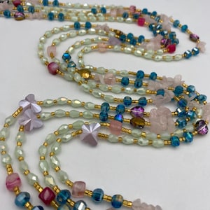 May include: A collection of layered necklaces featuring a variety of beads in shades of pink, blue, and green. The necklaces include faceted beads, small gold-colored spacers, and decorative flower-shaped charms. The necklaces are displayed against a white background.