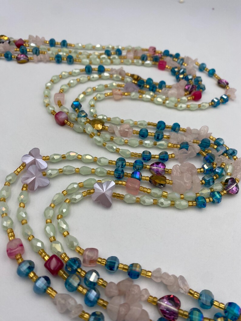 May include: A collection of layered necklaces featuring a variety of beads in shades of pink, blue, and green. The necklaces include faceted beads, small gold-colored spacers, and decorative flower-shaped charms. The necklaces are displayed against a white background.