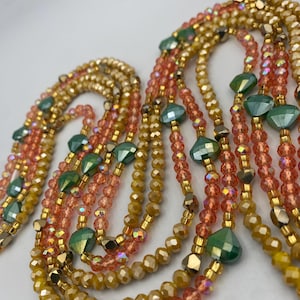 May include: Close-up of several beaded necklaces in shades of gold, peach, and green. The necklaces are made up of small, faceted beads, with larger, geometric green beads. The necklaces are arranged in a cascading design, creating a layered effect.