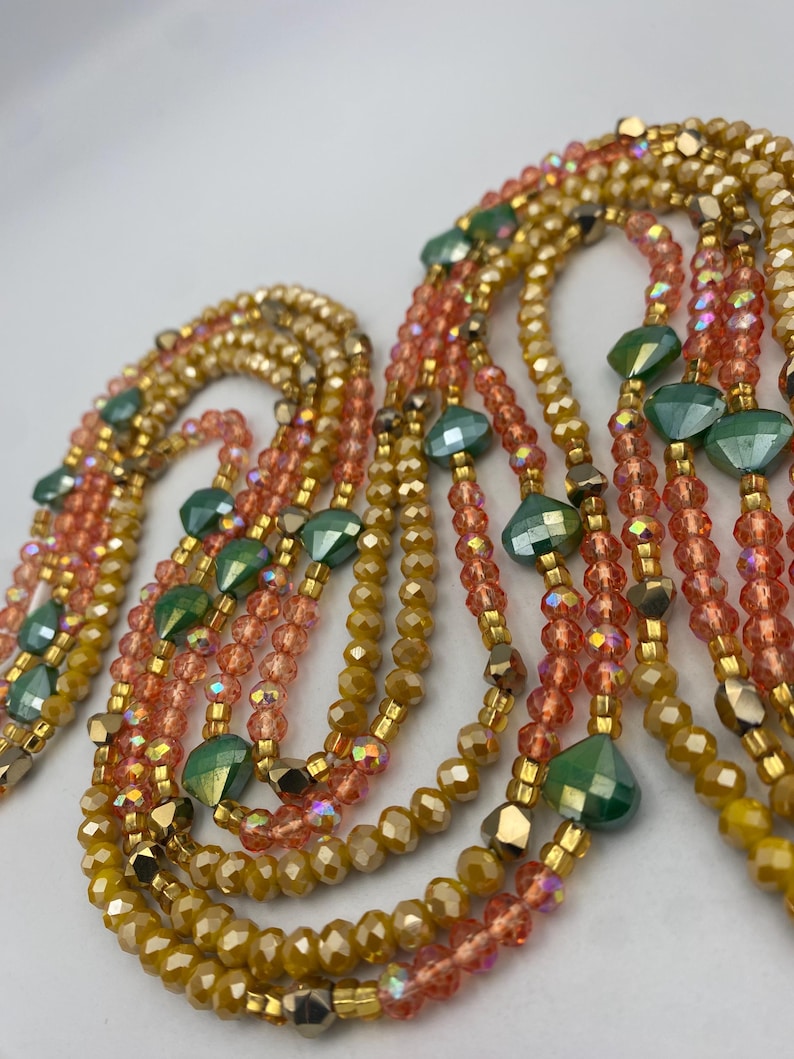 May include: Close-up of several beaded necklaces in shades of gold, peach, and green. The necklaces are made up of small, faceted beads, with larger, geometric green beads. The necklaces are arranged in a cascading design, creating a layered effect.