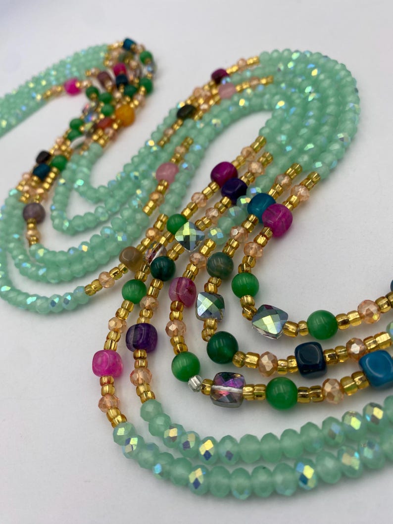 May include: Mint green beaded waist beads with gold accents and colourful beads. The strands feature a mix of round and faceted beads in shades of pink, green, purple, and blue. The beads are strung together with gold-coloured string.