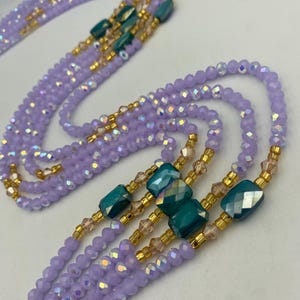 May include: Several strands of waist beads in lavender, teal, and gold. The beads are arranged in multiple strands, with teal rectangular beads and gold accents. The beads are small and faceted, creating a shimmering effect.