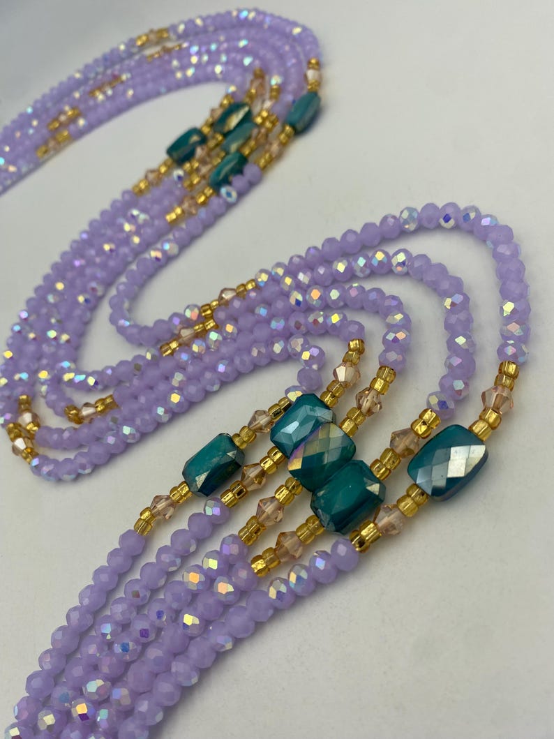 May include: Several strands of waist beads in lavender, teal, and gold. The beads are arranged in multiple strands, with teal rectangular beads and gold accents. The beads are small and faceted, creating a shimmering effect.