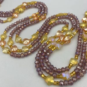 May include: Close-up of several beaded necklaces. The necklaces feature a mix of purple, yellow, and peach-coloured beads. Some beads are faceted, and others are shaped like flowers. The beads are strung on gold-coloured wire.