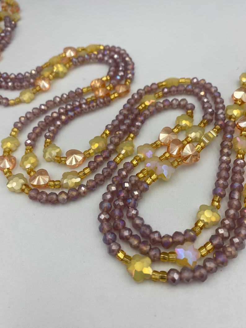 May include: Close-up of several beaded necklaces. The necklaces feature a mix of purple, yellow, and peach-coloured beads. Some beads are faceted, and others are shaped like flowers. The beads are strung on gold-coloured wire.