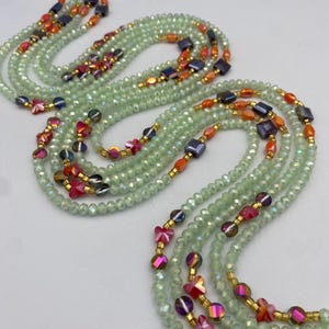 May include: Multiple strands of light green beaded jewellery with orange, red, and dark blue bead accents. The jewellery features small, faceted beads and gold-coloured spacers. The strands are arranged in a flowing, layered design.