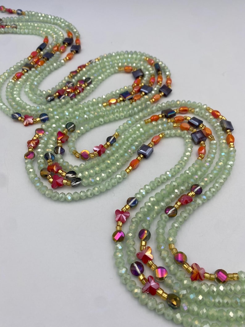 May include: Multiple strands of light green beaded jewellery with orange, red, and dark blue bead accents. The jewellery features small, faceted beads and gold-coloured spacers. The strands are arranged in a flowing, layered design.