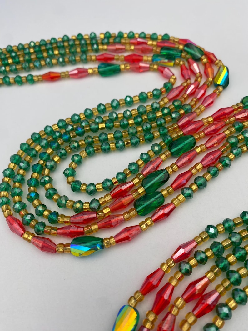 May include: A selection of beaded necklaces in green, red, and gold. The necklaces feature small, round beads and larger, faceted beads. The beads are strung together with gold-coloured string, creating a layered effect. The design is colourful and eye-catching.