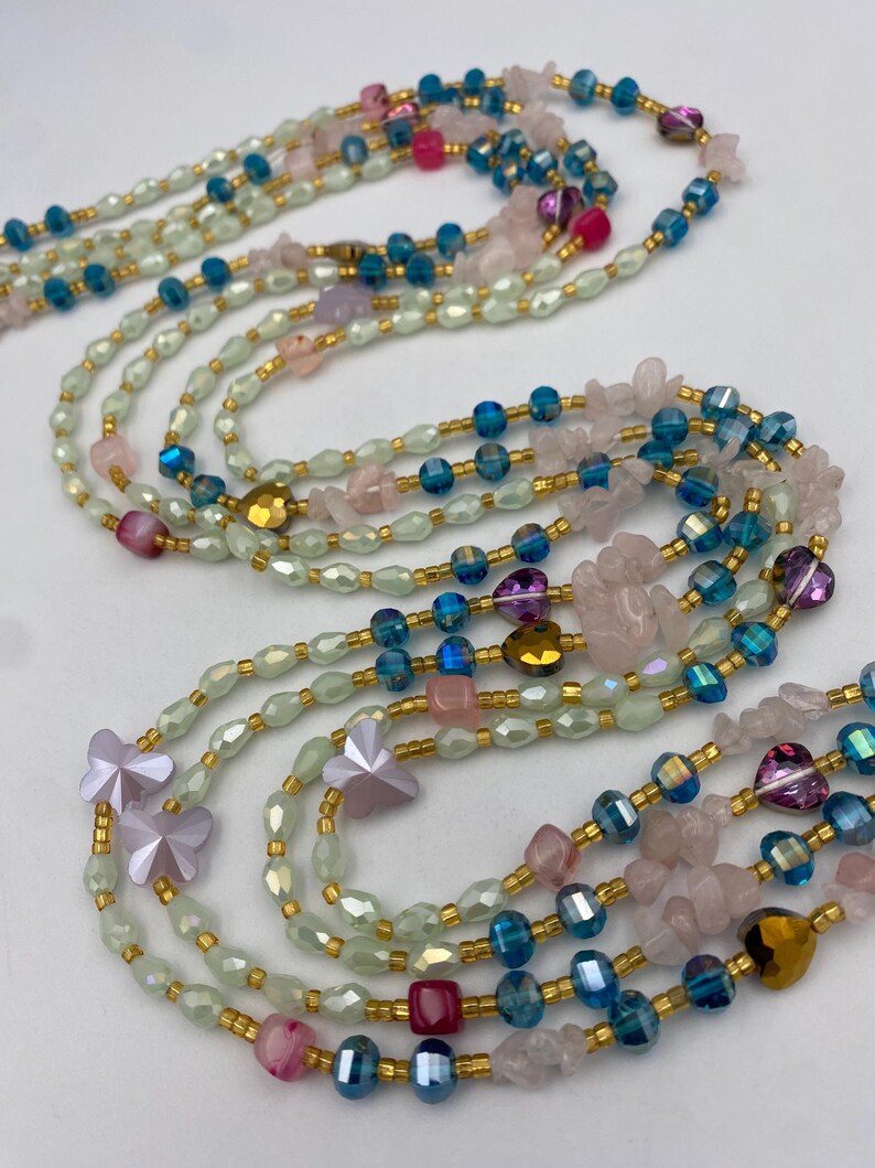 May include: Several layered necklaces with a variety of beads in shades of pink, blue, and green. The necklaces feature gold-colored accents and include butterfly and heart-shaped beads. The beads are strung together with gold-colored wire.