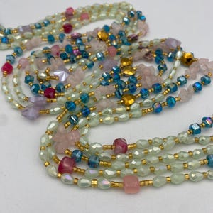 May include: A collection of beaded necklaces in various colors. The necklaces feature a mix of pink, blue, green, and gold beads. Some beads are iridescent, and others are shaped like squares or hearts. The necklaces are layered and appear to be handmade.