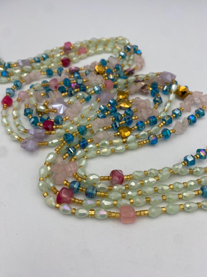 May include: A collection of beaded necklaces in various colors. The necklaces feature a mix of pink, blue, green, and gold beads. Some beads are iridescent, and others are shaped like squares or hearts. The necklaces are layered and appear to be handmade.